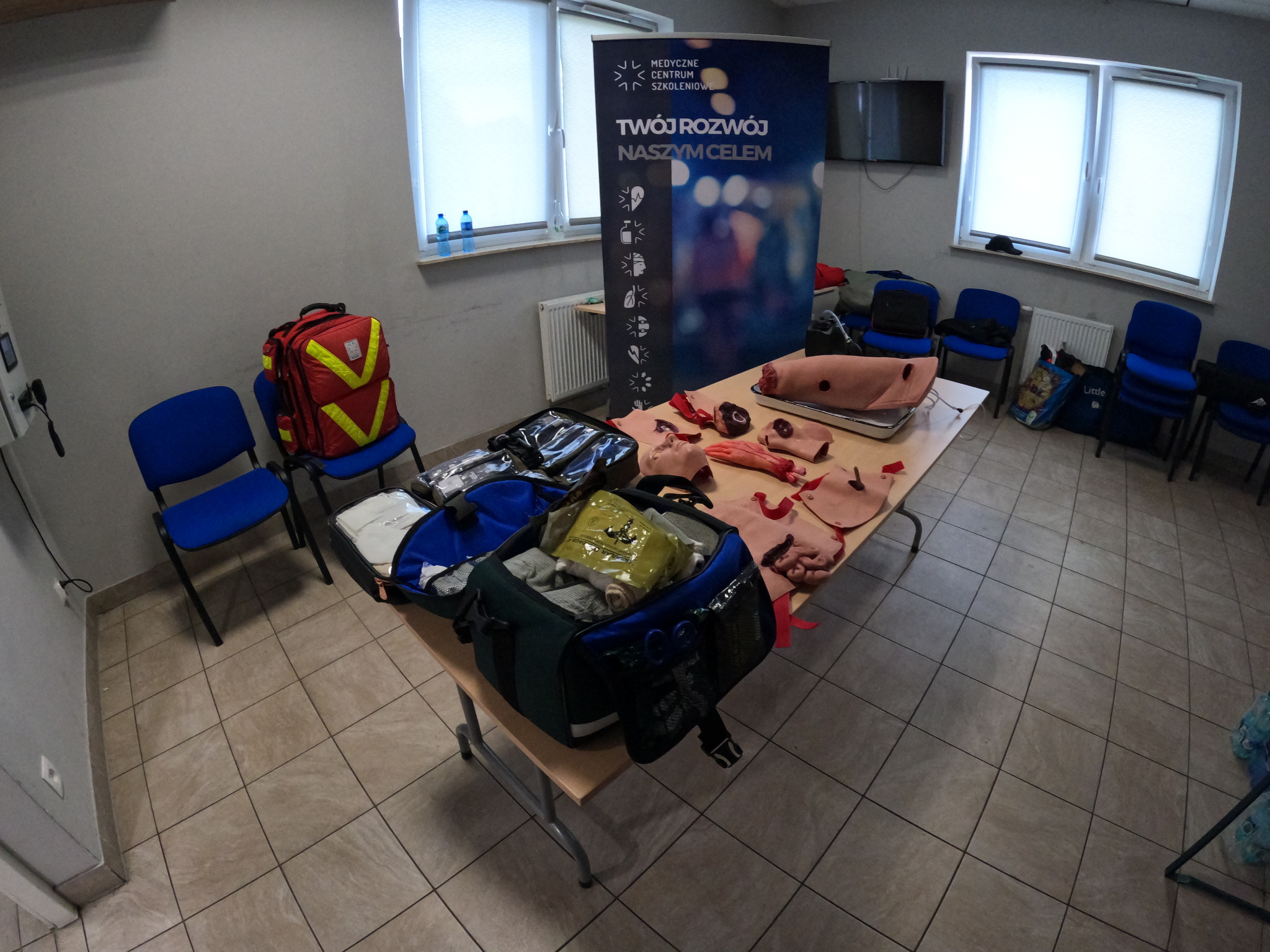 Sala szkoleniowa z fantomami do ćwiczeń pierwszej pomocy, torbami medycznymi i plecakiem ratunkowym na krzesłach.
