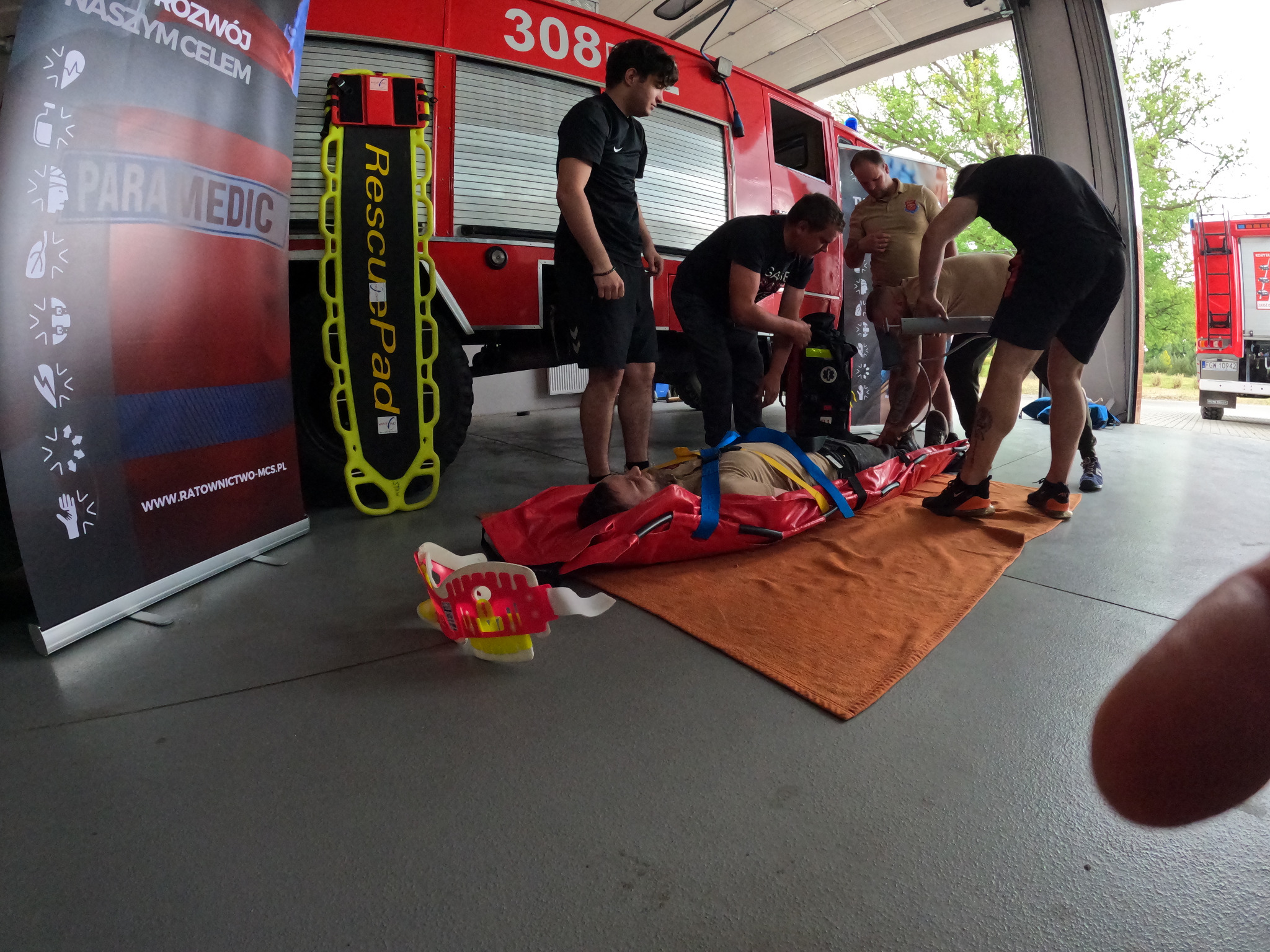 Symulacja akcji ratunkowej: mężczyzna leży na czerwonych noszach na pomarańczowym kocu, zabezpieczony niebieskimi pasami, otoczony przez ratowników w trakcie ćwiczeń, w tle wóz strażacki i baner...