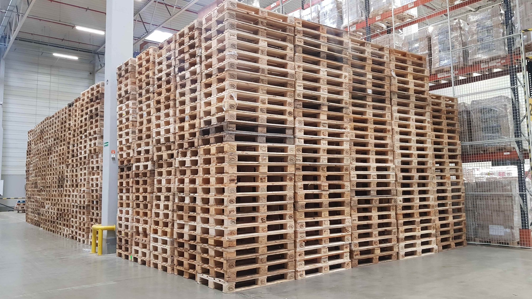 Wysoki stos drewnianych palet EPAL składowanych w magazynie, widoczne regały z zapakowanymi towarami w tle.