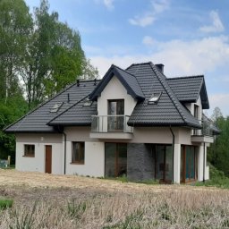 Extradom.pl - Nowoczesny dom z dwoma balkonami z przeszklonymi balustradami, ciemna dachówka, jasna elewacja z elementami dekoracyjnymi z kamienia, widok z poziomu gruntu na niezagospodarowaną działkę.
