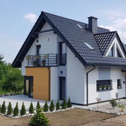 Extradom.pl - Nowoczesny dom z dachem z ciemnej dachówki, drewnianą elewacją i balkonem z kwiatami, otoczony trawnikiem z młodymi tujami.