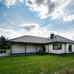 Parterowy dom z kremową elewacją i zadaszonym tarasem, widok z ogrodu w słoneczny dzień.