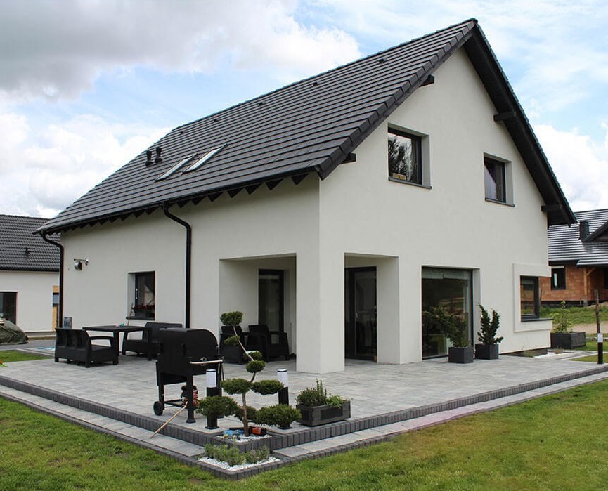 Nowoczesny dom murowany z białym tynkiem, ciemną dachówką i tarasem z szarej kostki, wyposażonym w grill i meble ogrodowe, otoczony zielonym trawnikiem.