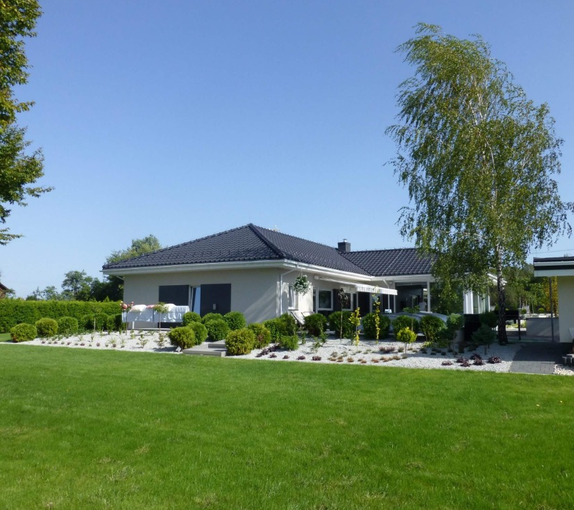 Parterowy dom z ciemnym dachem, kremową elewacją i starannie zagospodarowanym ogrodem z białym żwirem i niskimi krzewami.