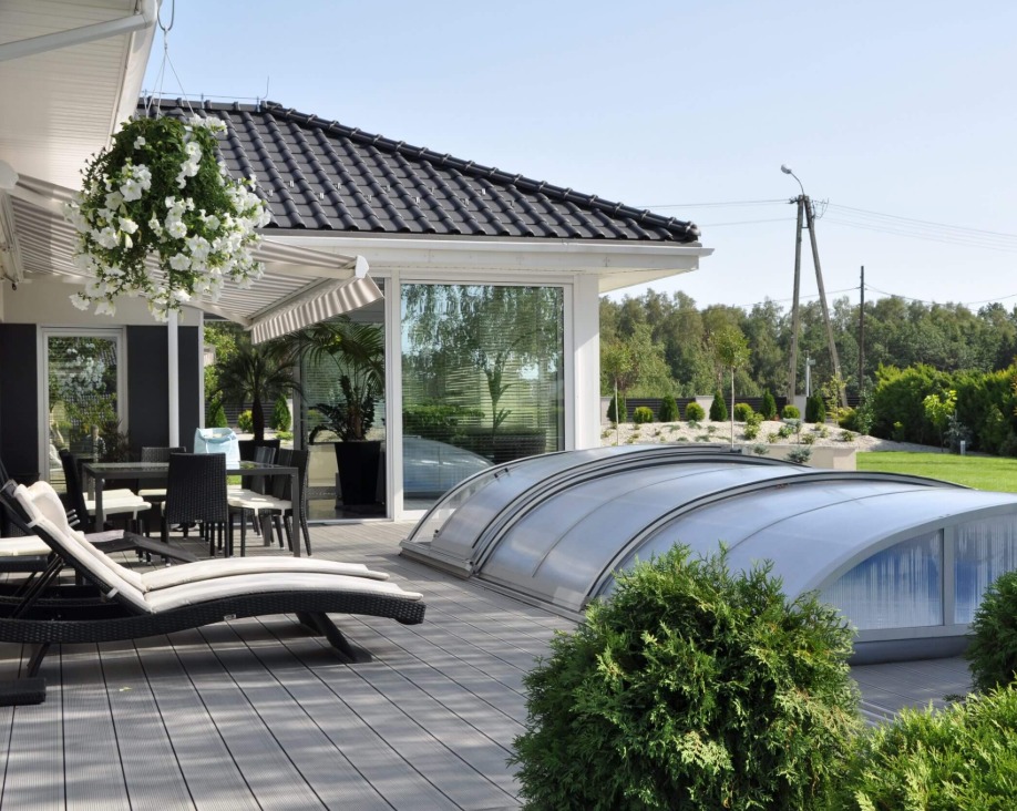 Nowoczesny dom z tarasem z szarej deski kompozytowej, zadaszeniem basenu i wiszącą donicą z białymi kwiatami.