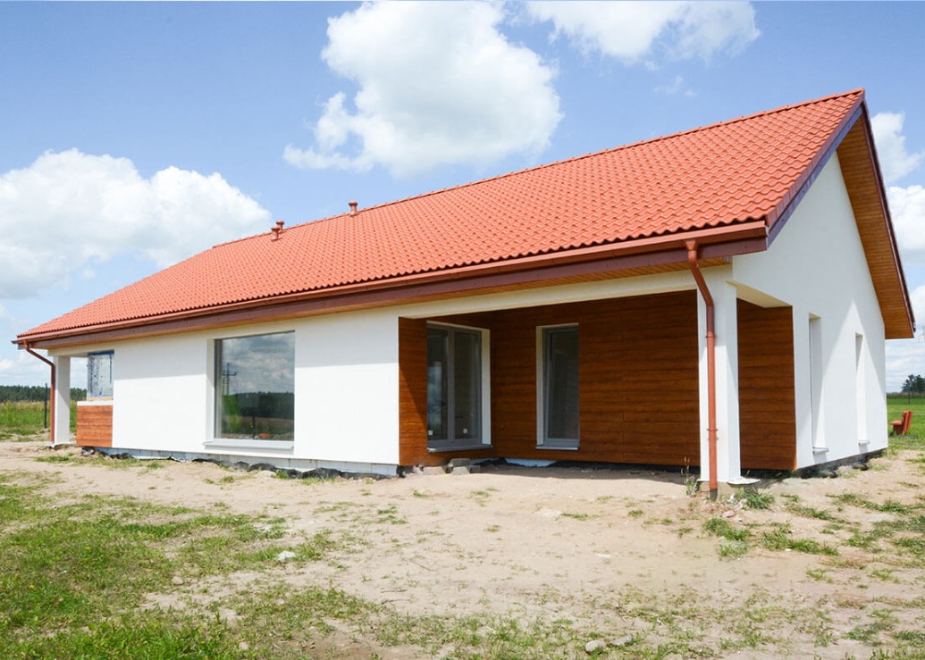 Parterowy dom z czerwoną dachówką i drewnianą elewacją, częściowo otynkowany na biało, z widocznym fragmentem fundamentu i niezagospodarowanym terenem wokół.