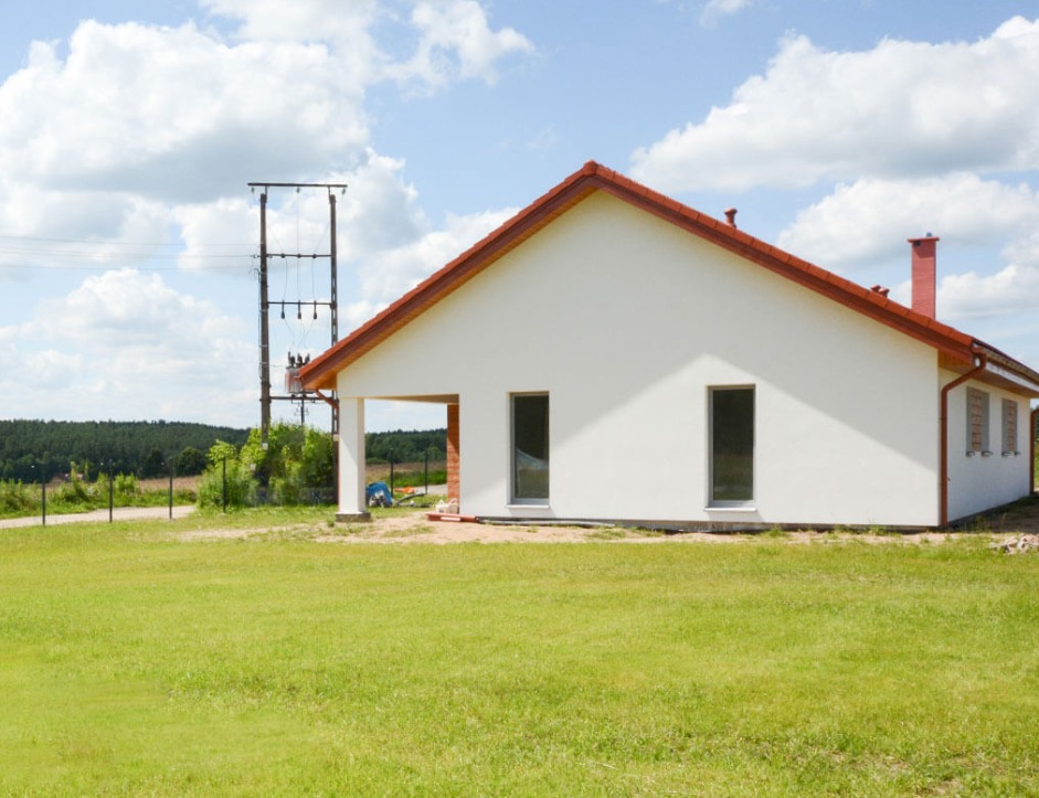 Biały parterowy dom z czerwonym dachem i kominem, widok na trawnik i słup wysokiego napięcia w tle, częściowo wykończony.