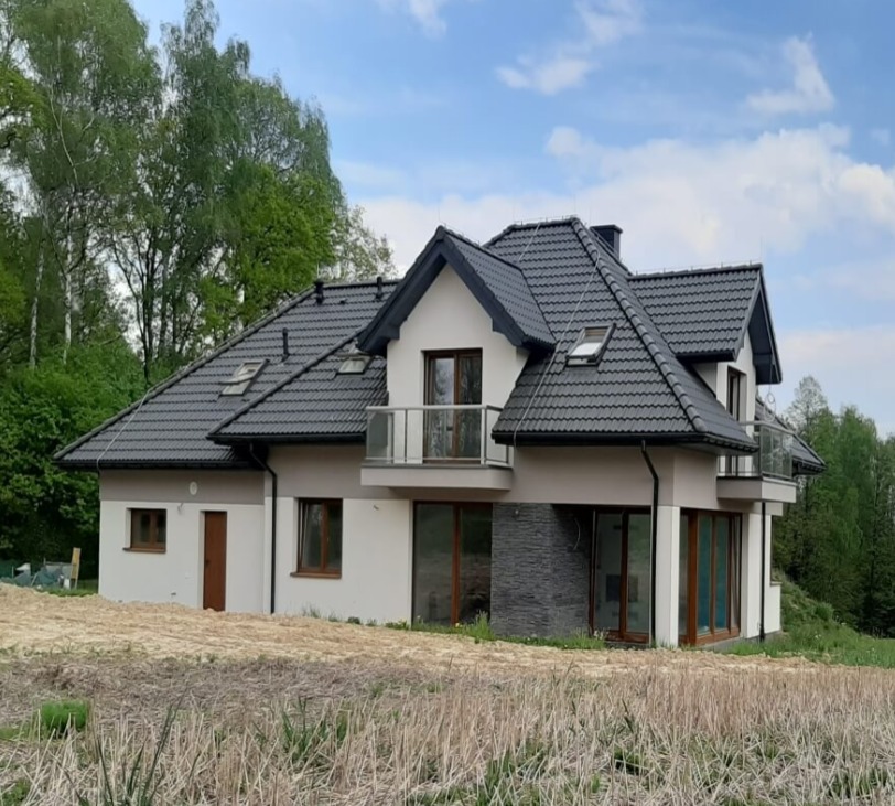 Nowoczesny dom z dwoma balkonami z przeszklonymi balustradami, ciemna dachówka, jasna elewacja z elementami dekoracyjnymi z kamienia, widok z poziomu gruntu na niezagospodarowaną działkę.