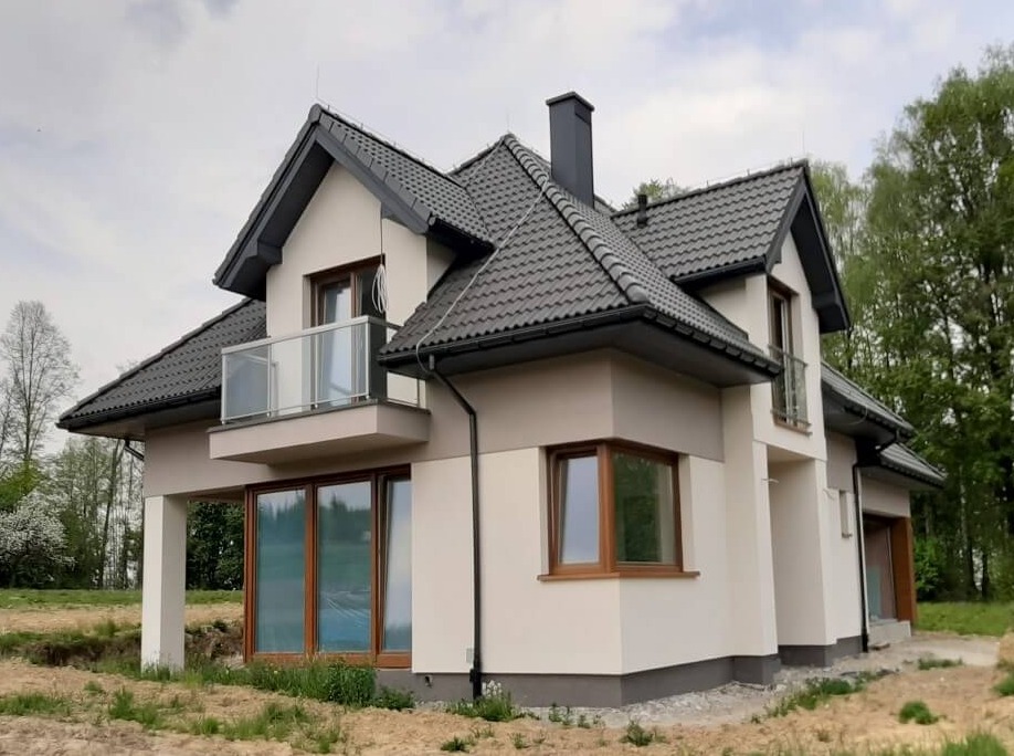 Nowoczesny dom z balkonem z szklaną balustradą, ciemna dachówka i jasna elewacja, widoczne okna z drewnianymi ramami, otoczenie w trakcie zagospodarowywania.