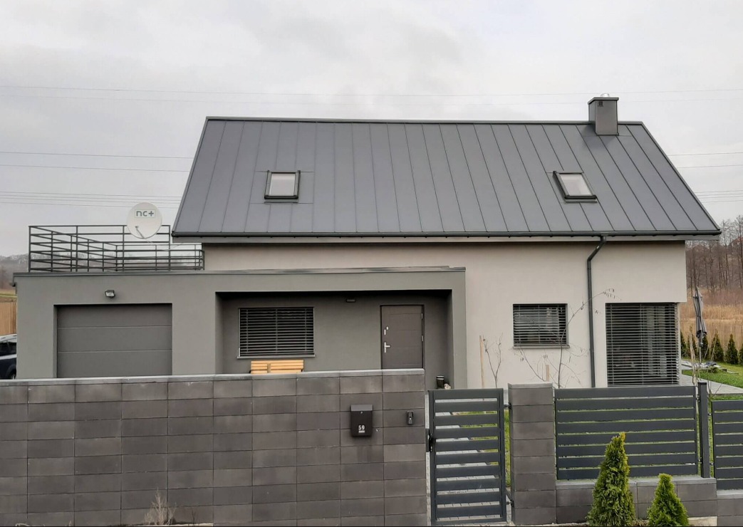 Nowoczesny dom jednorodzinny z szarą, metalową elewacją i dachem, widoczny zza ogrodzenia z betonowych bloczków. Na dachu satelita i dwa okna dachowe.