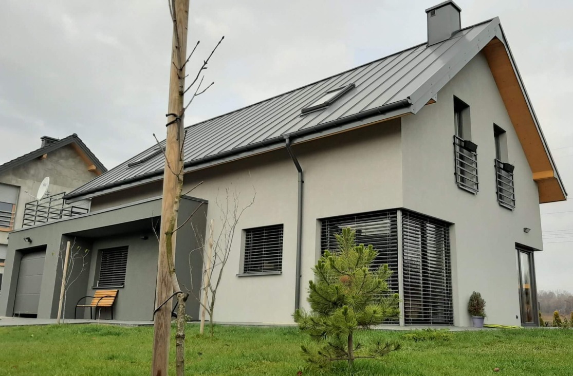 Nowoczesny dom jednorodzinny z szarą blachą na dachu, jasną elewacją i grafitowymi detalami, z widocznym fragmentem ogrodu i drzewkiem iglastym na pierwszym planie.