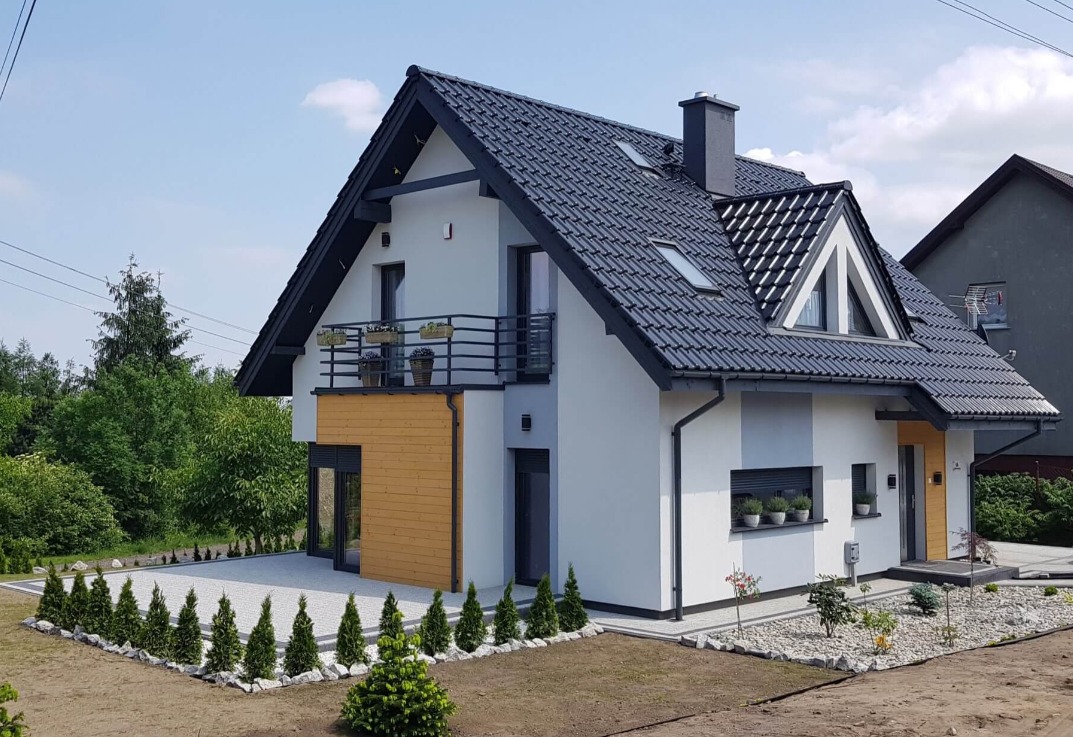 Nowoczesny dom z dachem z ciemnej dachówki, drewnianą elewacją i balkonem z kwiatami, otoczony trawnikiem z młodymi tujami.