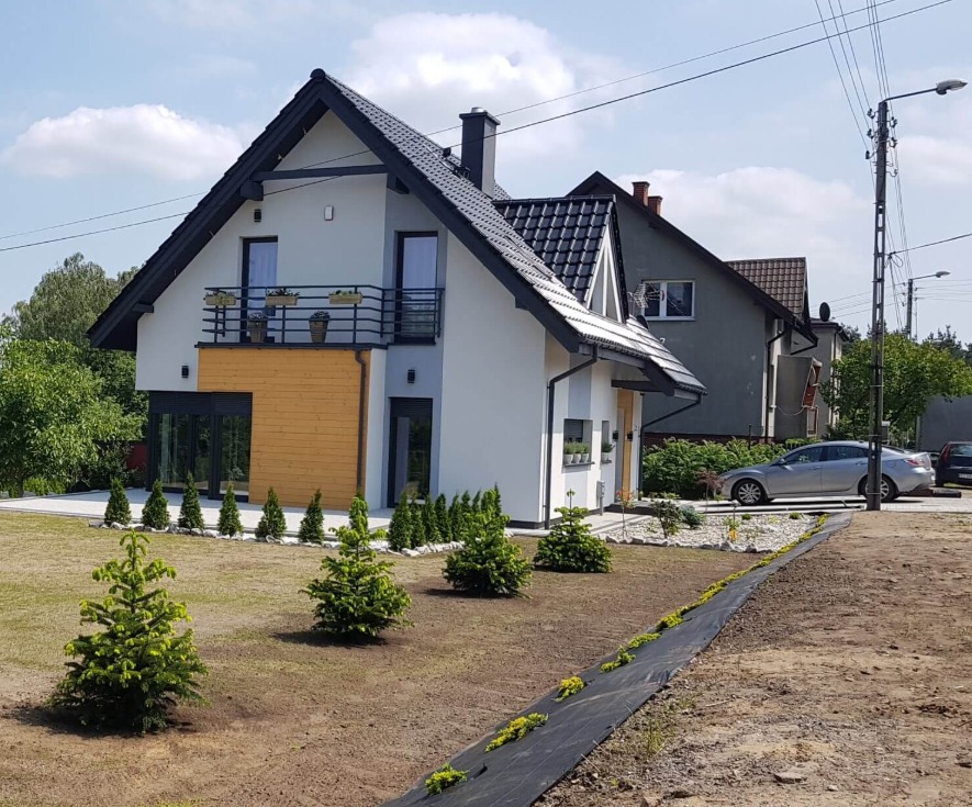 Nowoczesny dom z białymi ścianami, drewnianą elewacją i czarnym dachem, balkon z kwiatami, świeżo założony trawnik z młodymi drzewkami iglastymi.