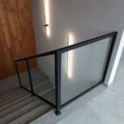 Nowoczesna szklana balustrada z czarną ramą na schodach wewnątrz budynku, oświetlona pionowymi lampami ściennymi, z drewnianą ścianą w tle.