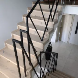 Schody z jasnego drewna z czarną, geometryczną balustradą, widok z góry, z doniczką z rośliną u podnóża schodów i drzwiami w tle.