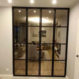 Szklane drzwi w czarnej metalowej ramie oddzielające pokój dzienny z choinką i beżową sofą od korytarza z szarą podłogą.