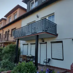 Nowoczesna, pozioma, metalowa balustrada balkonowa w kolorze antracytowym, na tle białej elewacji budynku z roletami zewnętrznymi i zadbanym ogrodem.