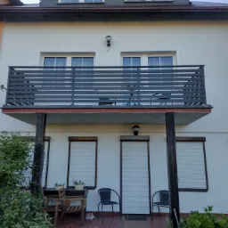 Biały dom z balkonem zabezpieczonym nowoczesną, poziomą, szarą konstrukcją; na balkonie widoczne krzesła i stolik.