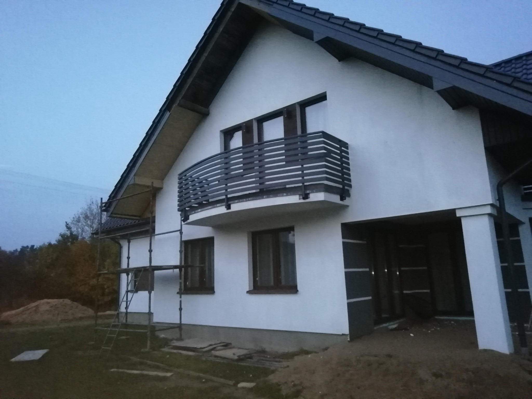 Dom jednorodzinny z białymi ścianami i balkonem z poziomymi, ciemnymi barierkami. Na pierwszym planie rusztowanie i usypana ziemia.