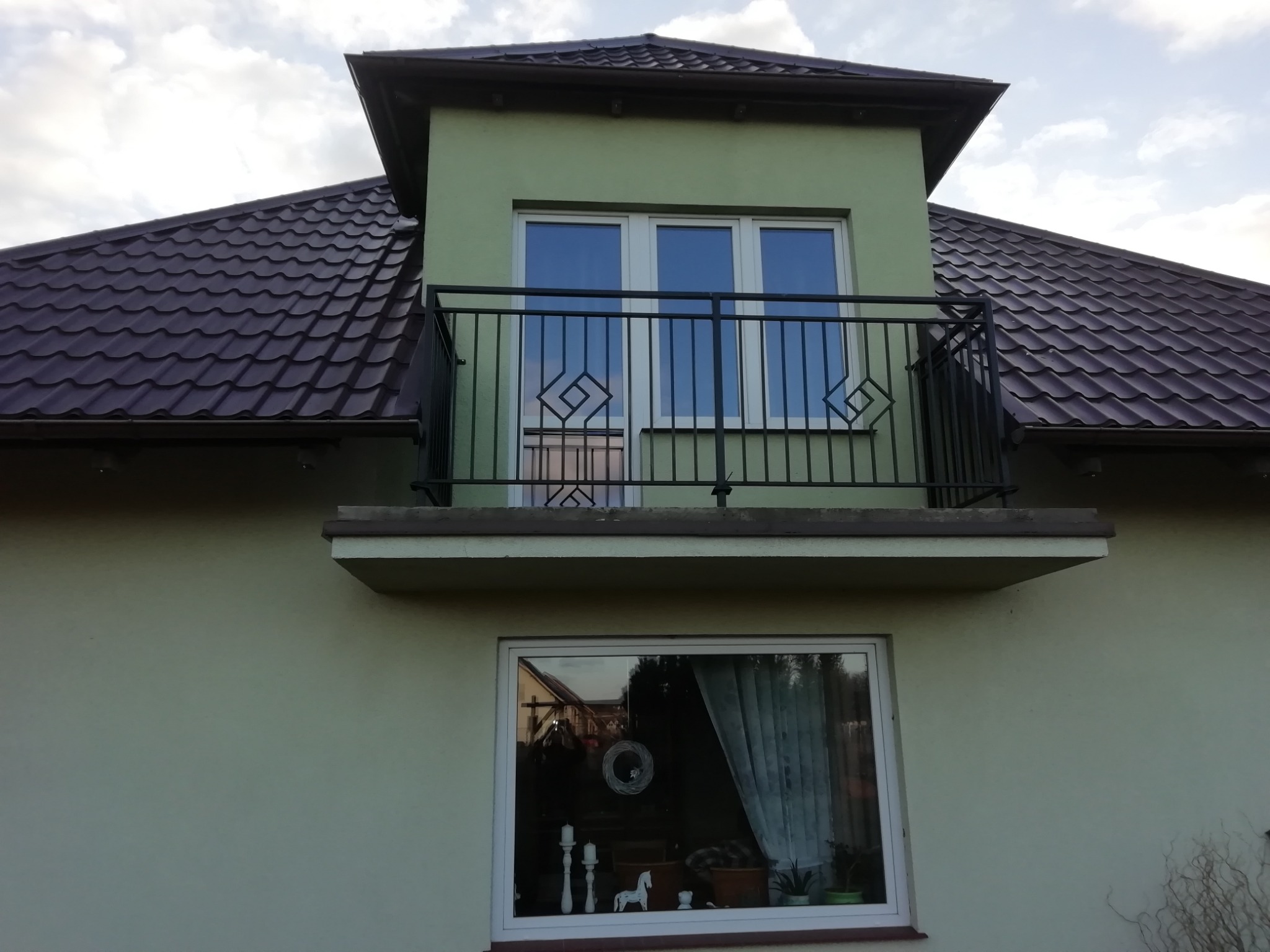 Balkon z czarną metalową balustradą o geometrycznym wzorze na tle zielonej elewacji domu z brązowym dachem.