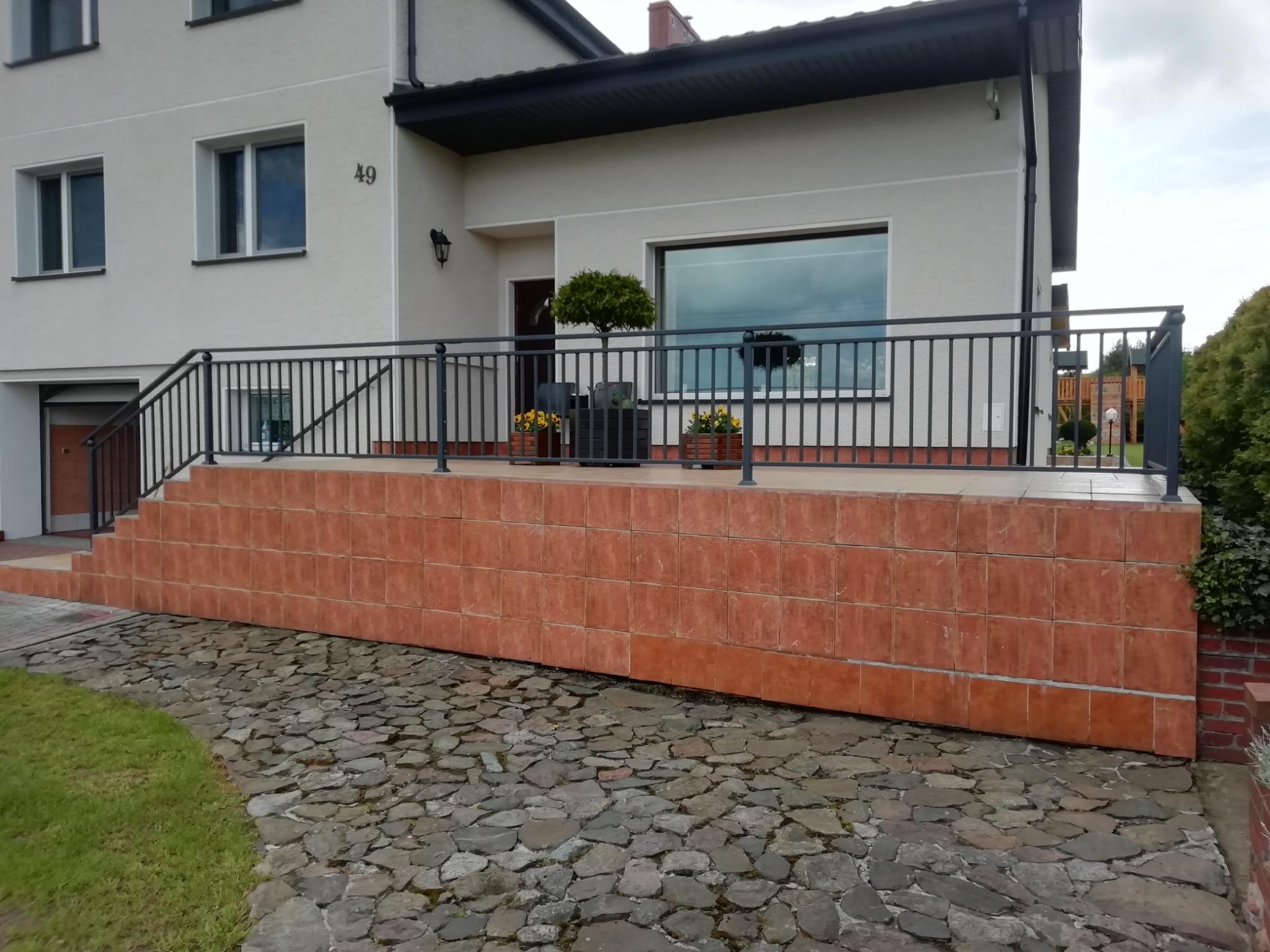 Ciemna, metalowa barierka z pionowymi szczeblami, zabezpieczająca taras przed domem z elewacją w kolorze złamanej bieli i numerem 49, widok z poziomu brukowanej ścieżki.