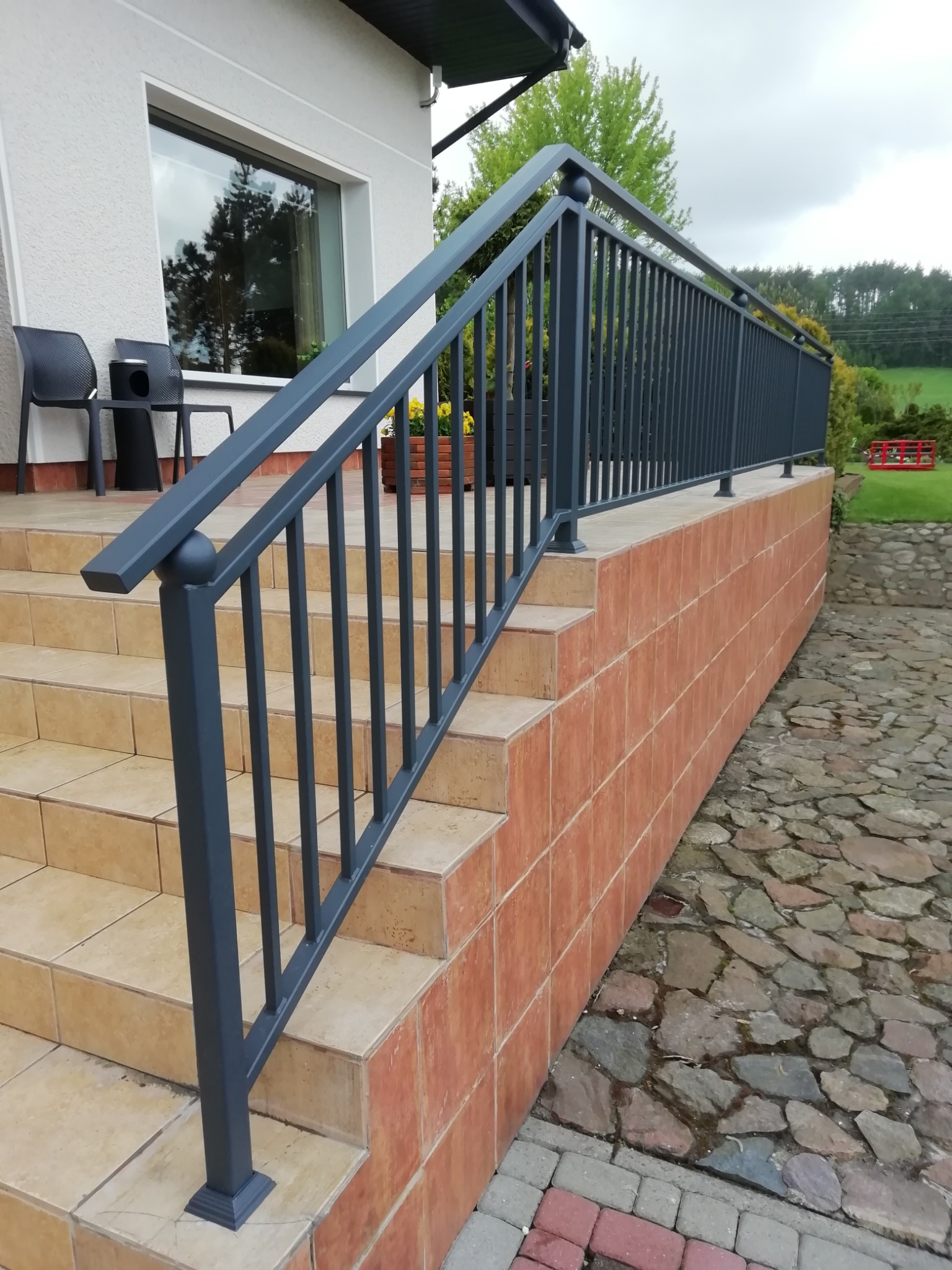 Zewnętrzne schody z beżowymi płytkami i szarą, metalową balustradą z kulistymi zakończeniami, obok ściana wyłożona kafelkami w kolorze terakoty i kamienny chodnik