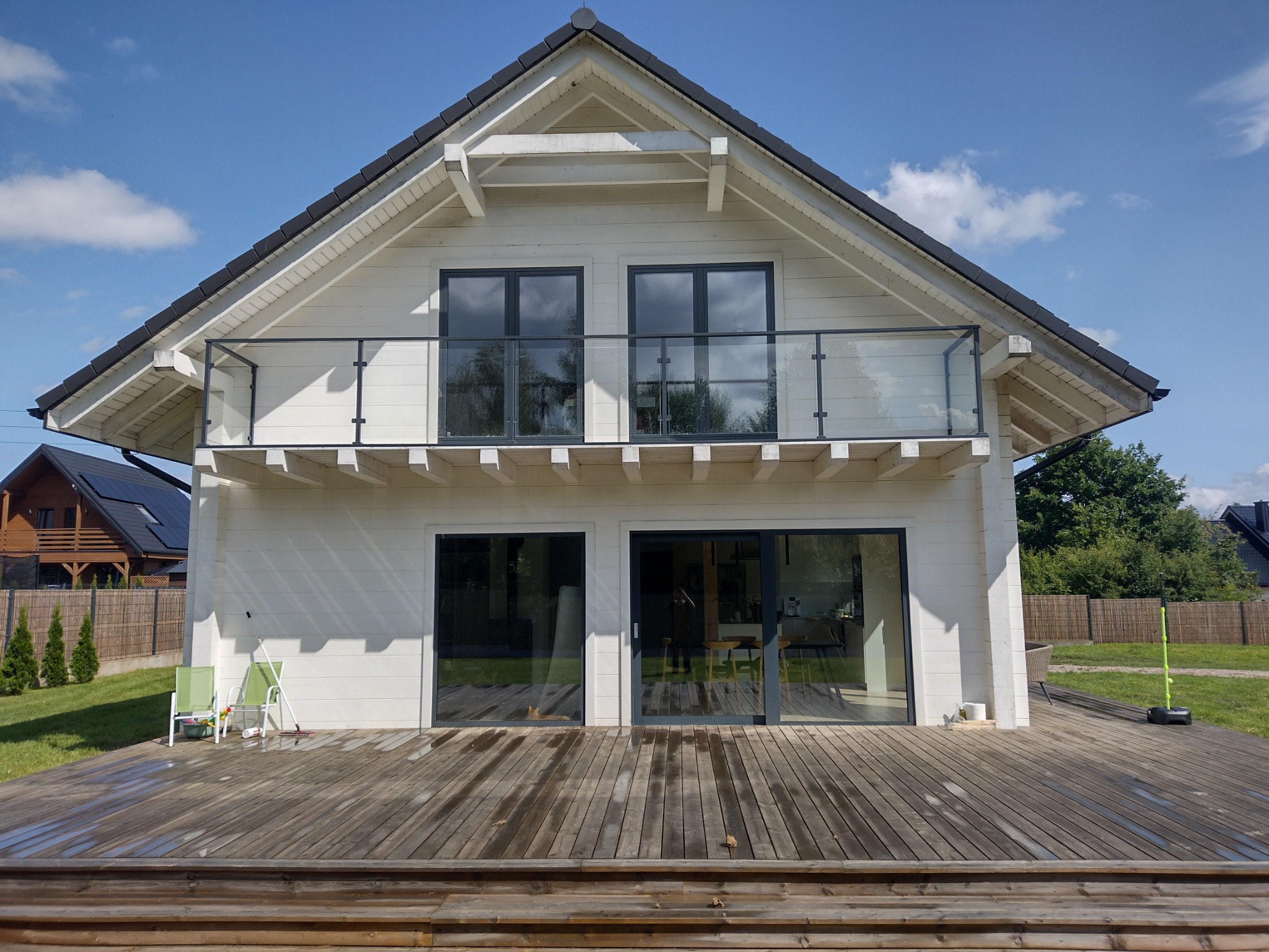 Nowoczesny, biały dom z drewnianą elewacją, dużym tarasem i balkonem zabezpieczonym szklaną taflą, widok z dołu na tle błękitnego nieba.