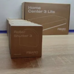Stos kartonowych pudełek Fibaro Roller Shutter 3 na drewnianym blacie, w tle pudełko Fibaro Home Center 3 Lite.