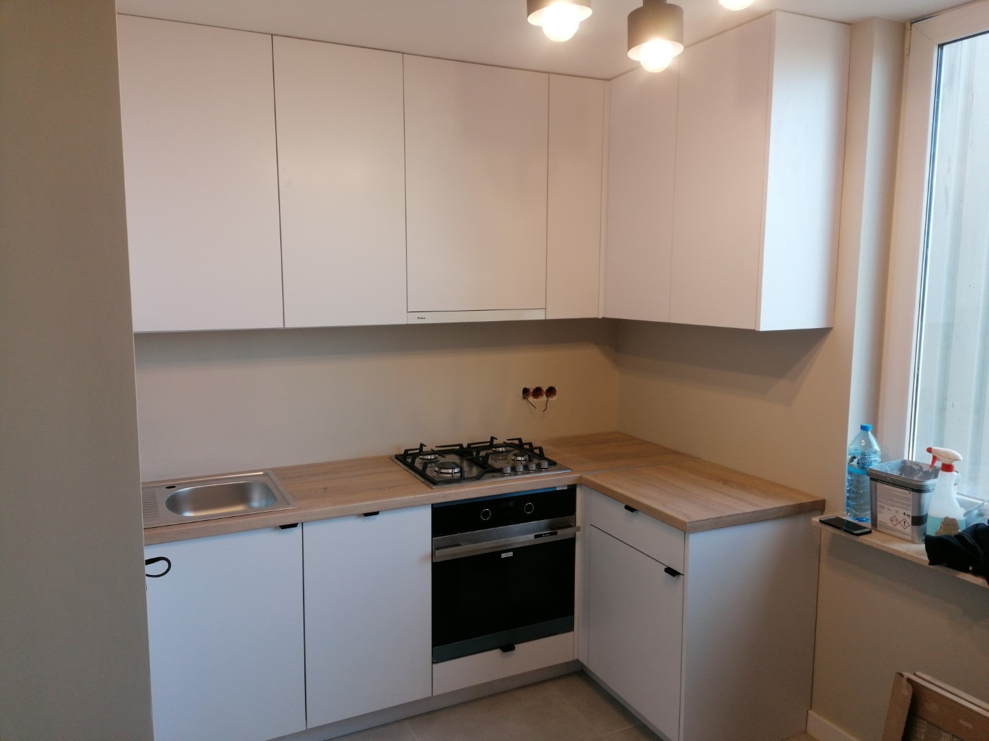 Biała kuchnia na wymiar z drewnianym blatem, widoczne gniazdka elektryczne nad kuchenką, minimalistyczny styl.