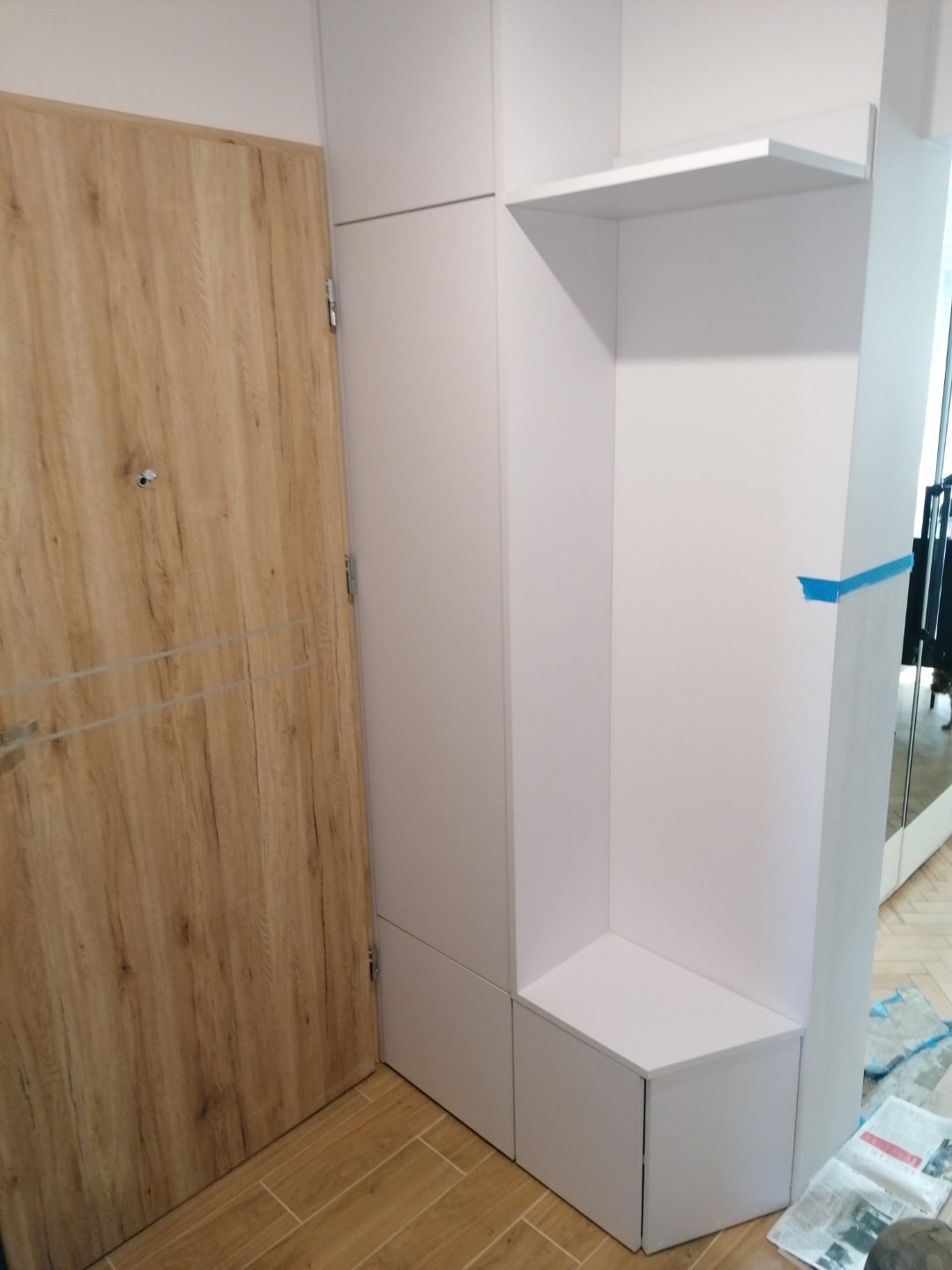 Biała zabudowa meblowa z półką i siedziskiem przy drewnianych drzwiach wejściowych, widoczne gazety na podłodze.