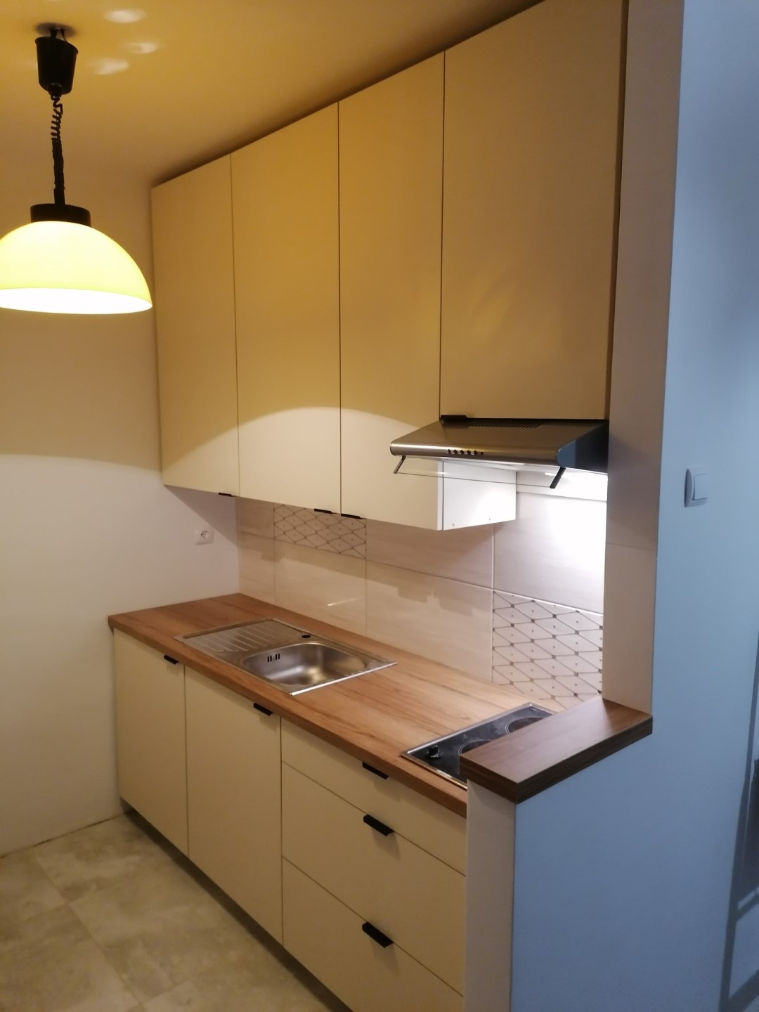 Jasna zabudowa kuchenna z drewnianym blatem, czarnymi uchwytami i okapem, oświetlona wiszącą lampą z żółtym kloszem.