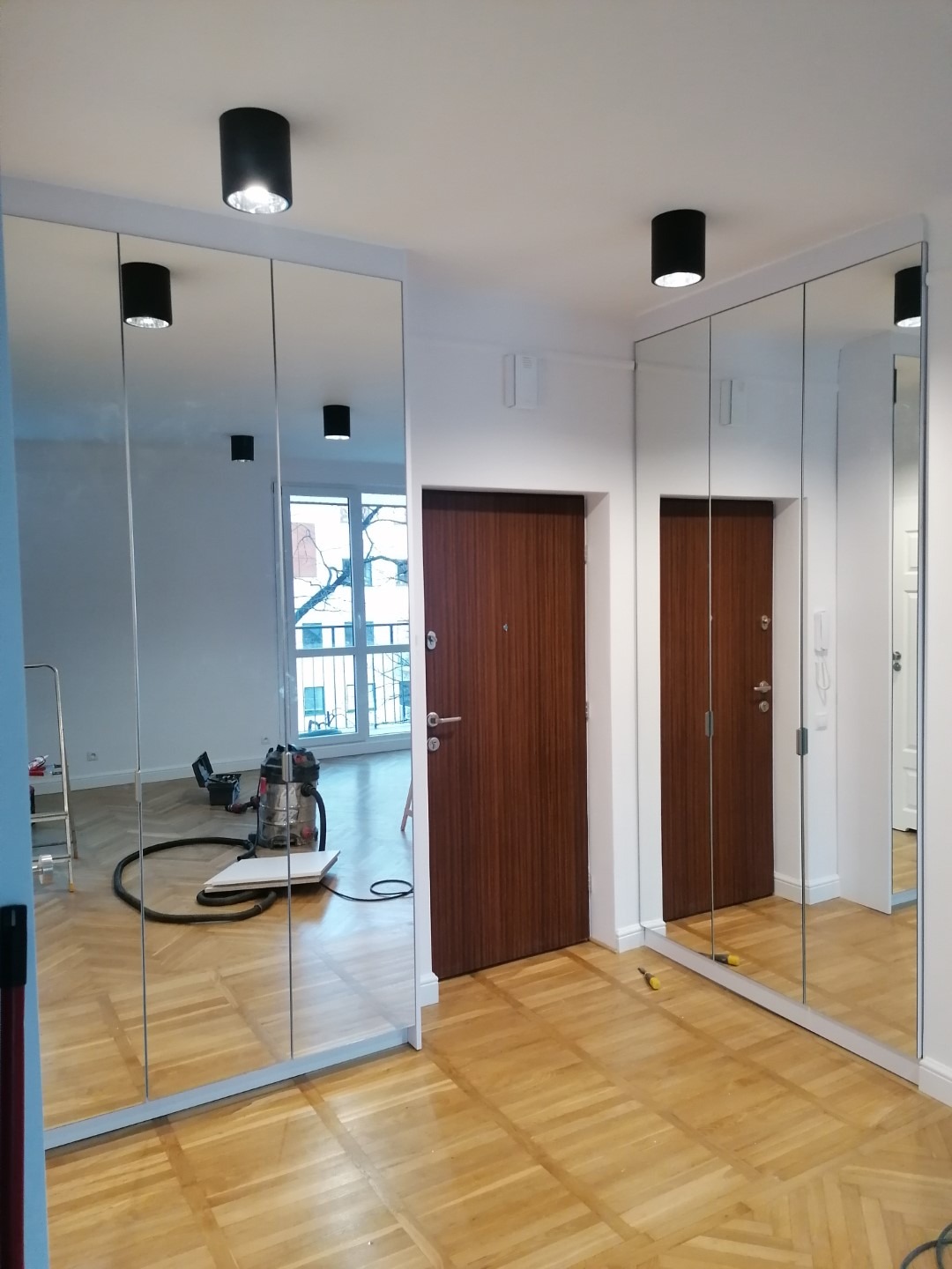 Korytarz z dwiema szafami z lustrzanymi drzwiami, widoczny parkiet i drzwi wejściowe, na podłodze leży śrubokręt, widoczny odkurzacz i drabina.