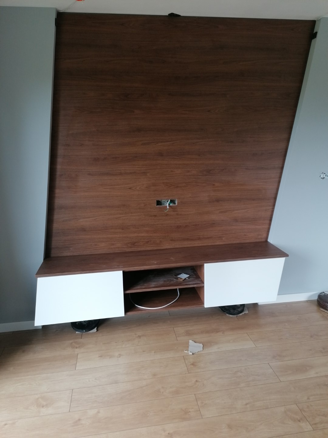 Montaż mebli RTV na ścianie z drewnianą okładziną, widoczne kable i elementy mocujące, podłoga w panelach.