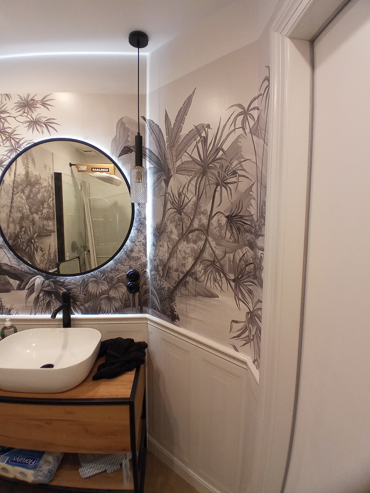 Umywalka nablatowa z czarną armaturą na drewnianej szafce w łazience z tapetą w motywy tropikalne i okrągłym lustrem z podświetleniem LED
