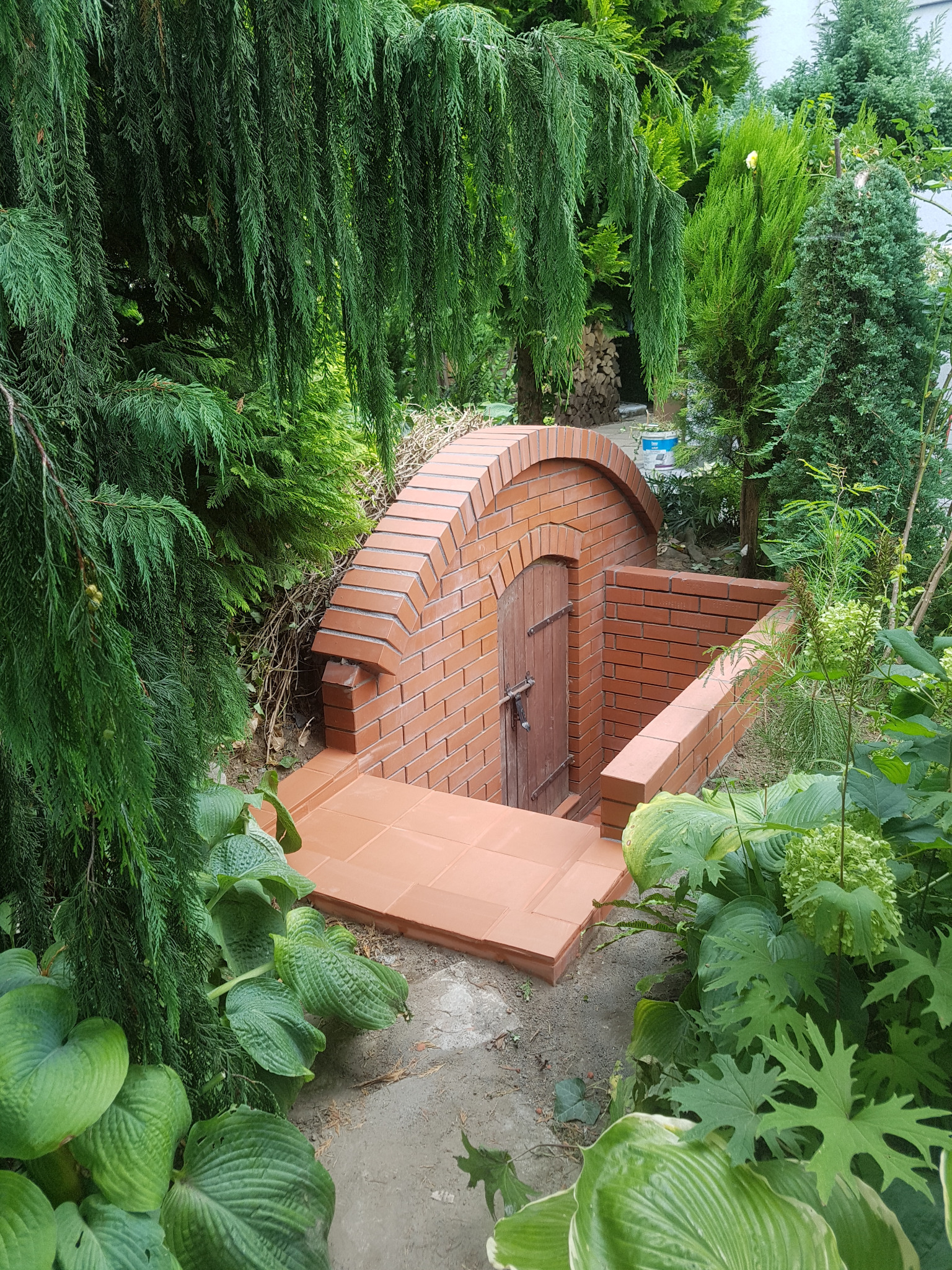 Murowana piwniczka ziemna z cegły z drewnianymi drzwiami, otoczona bujną roślinnością ogrodową