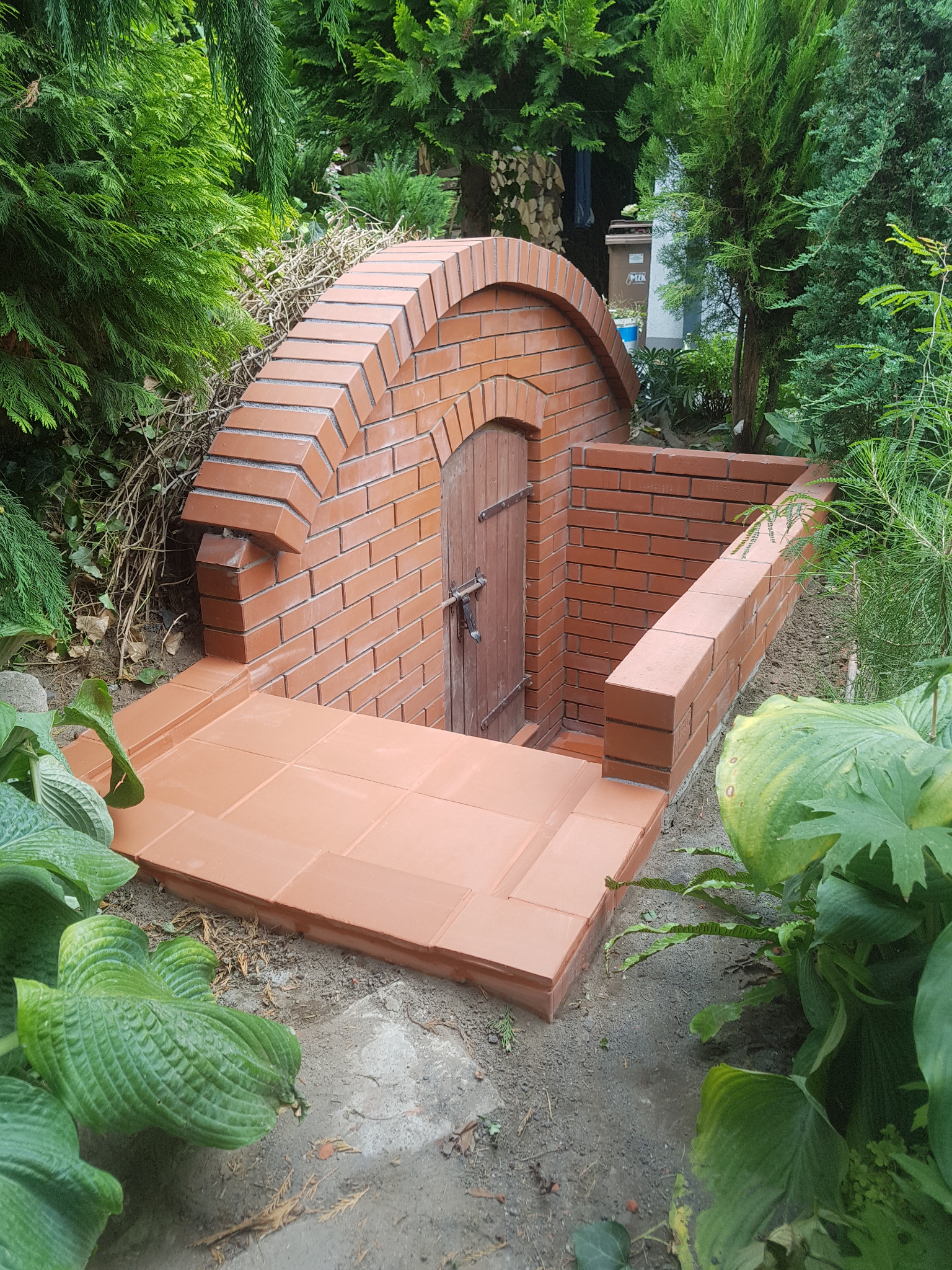 Zewnętrzne wejście do piwnicy z cegły z drewnianymi drzwiami, otoczone zielenią.