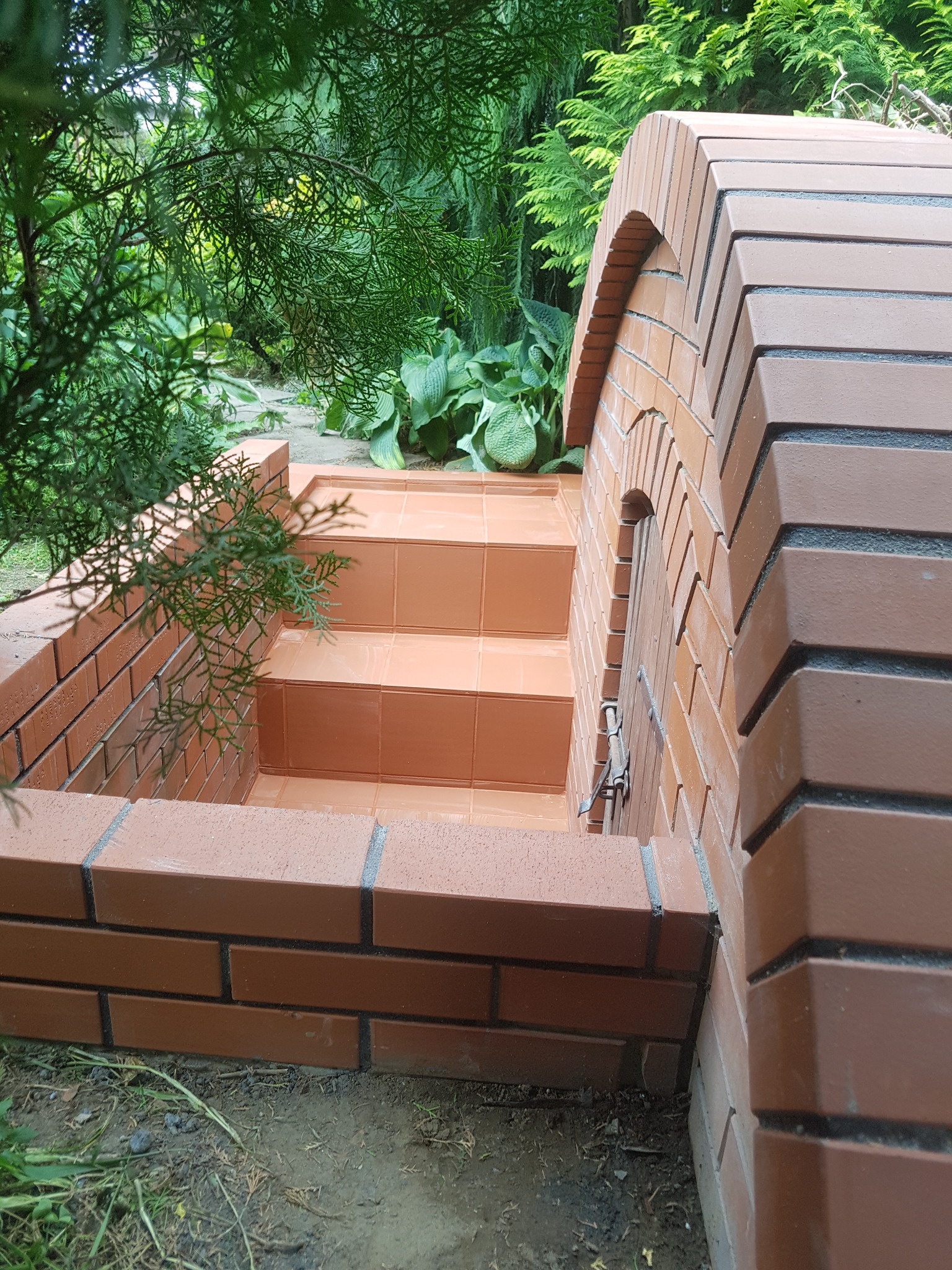 Murowana piwniczka ogrodowa z cegły klinkierowej, wyłożona terakotą, otoczona zielenią.