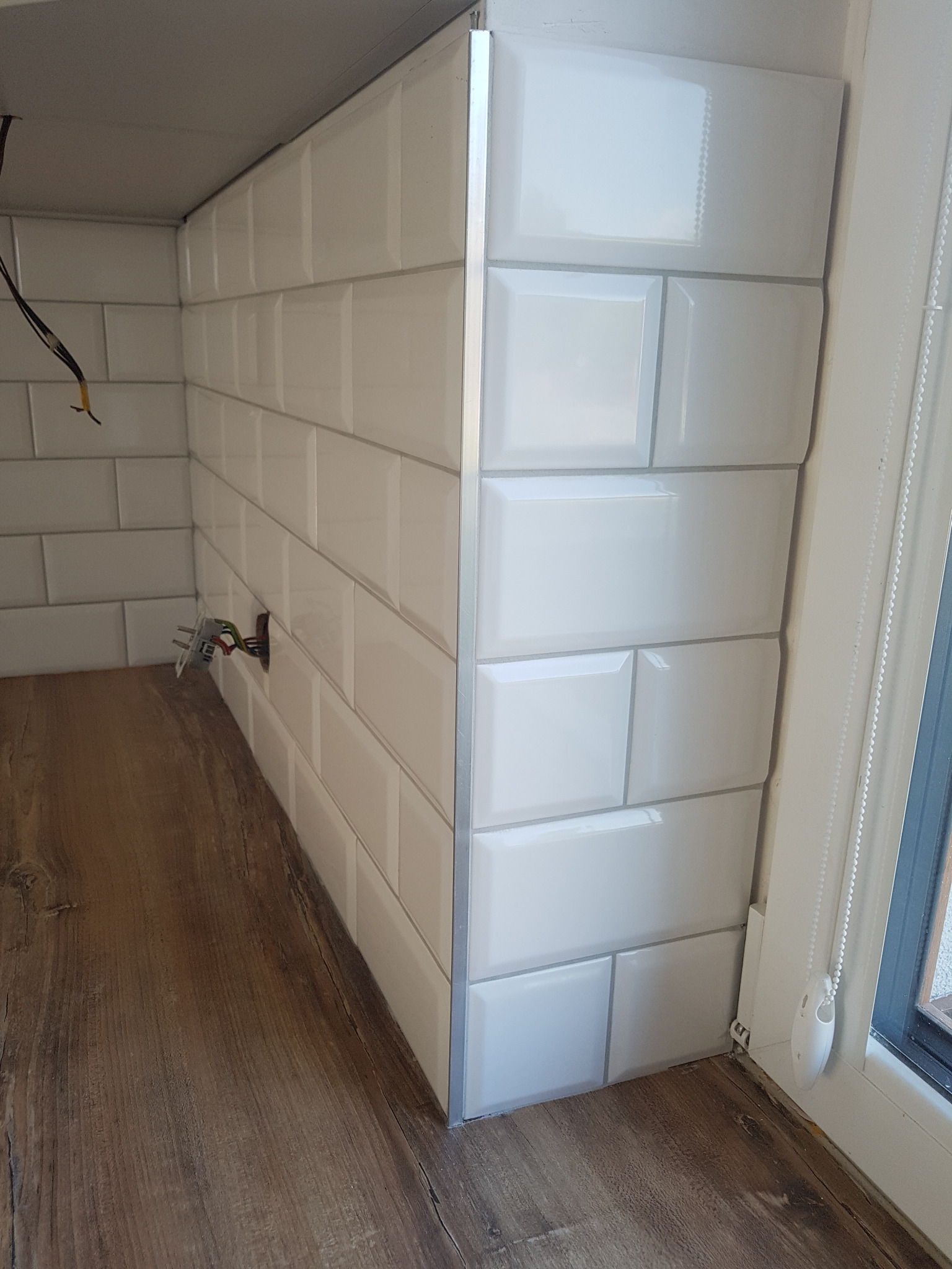 Białe, błyszczące płytki typu metro z szarą fugą na ścianie w trakcie układania, widoczne wystające przewody elektryczne i fragment drewnianego blatu w kuchni w Warszawie.