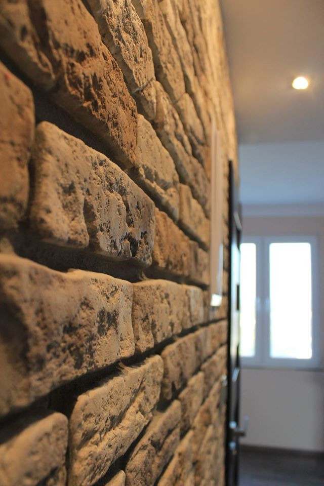 Ściana wykończona dekoracyjną cegłą w odcieniach beżu i brązu, widok z boku, w tle widoczne drzwi i okno z białymi ramami.