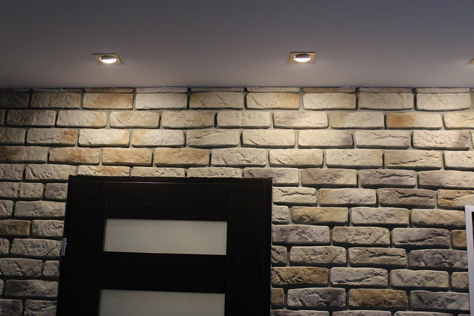 Ściana wykończona dekoracyjną cegłą w odcieniach beżu i szarości, oświetlona punktowymi lampami sufitowymi, z fragmentem ciemnych drzwi z poziomymi przeszkleniami.