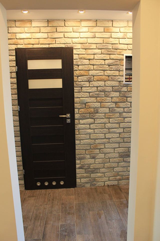 Ciemne drzwi wewnętrzne z poziomymi przeszkleniami, zamontowane w ścianie wyłożonej dekoracyjną cegłą w odcieniach beżu i szarości, na podłodze imitacja drewna.