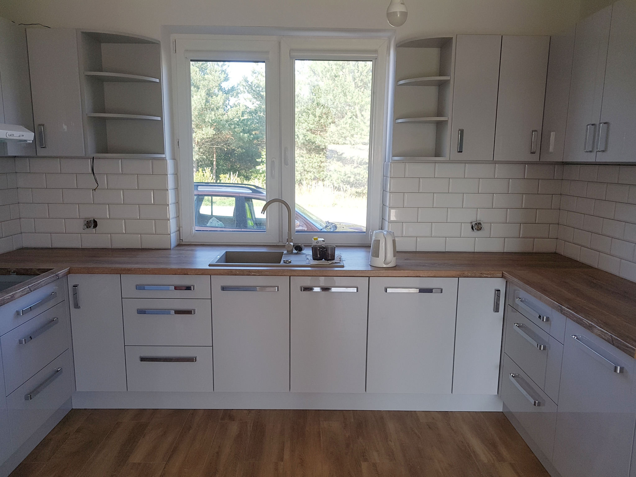 Jasna kuchnia z drewnianym blatem, białymi płytkami na ścianie i widokiem na zieleń za oknem