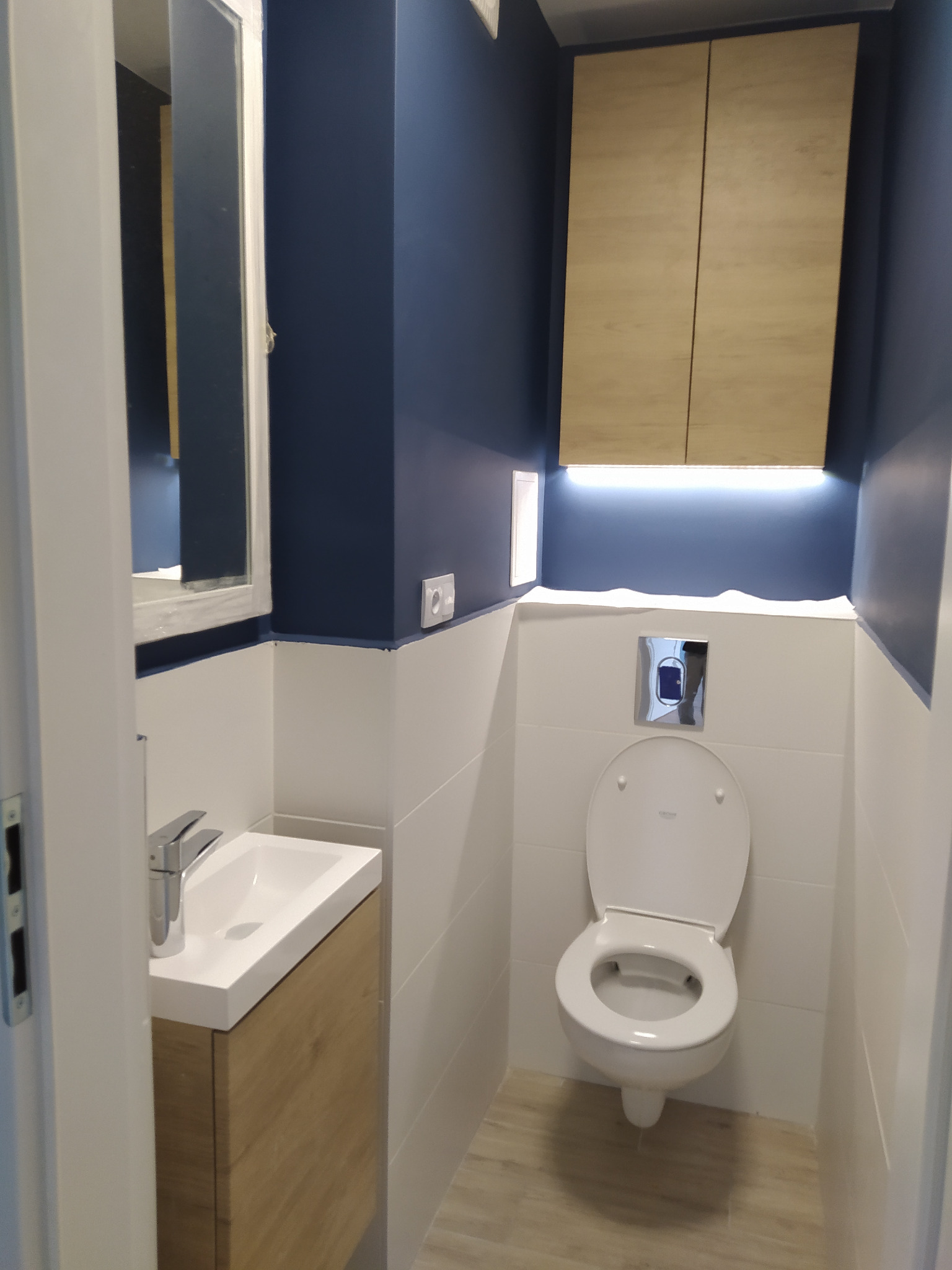 Nowoczesna łazienka z toaletą, umywalką z szafką z drewnopodobnym fornirem, szafką wiszącą i niebieskimi ścianami w górnej części.