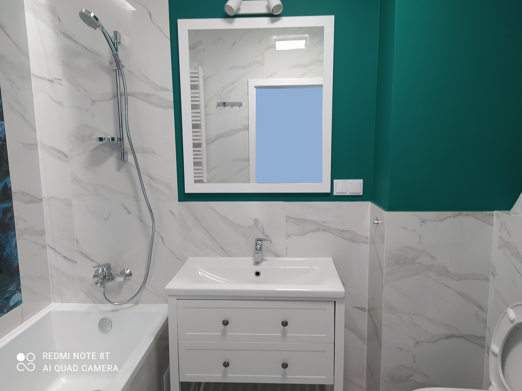 Nowoczesna łazienka z białymi marmurowymi płytkami, umywalką z szafką w stylu angielskim, lustrem z oświetleniem i akcentem w kolorze butelkowej zieleni na ścianie.