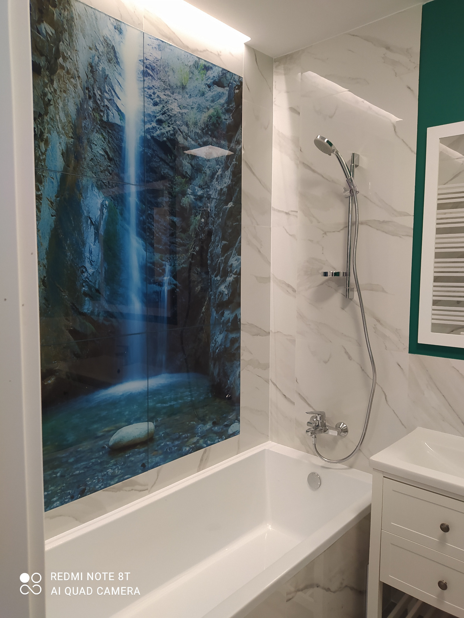 Nowoczesna łazienka z wanną obudowaną płytkami z motywem wodospadu, marmurowe płytki ścienne, chromowana armatura i biała szafka pod umywalką.