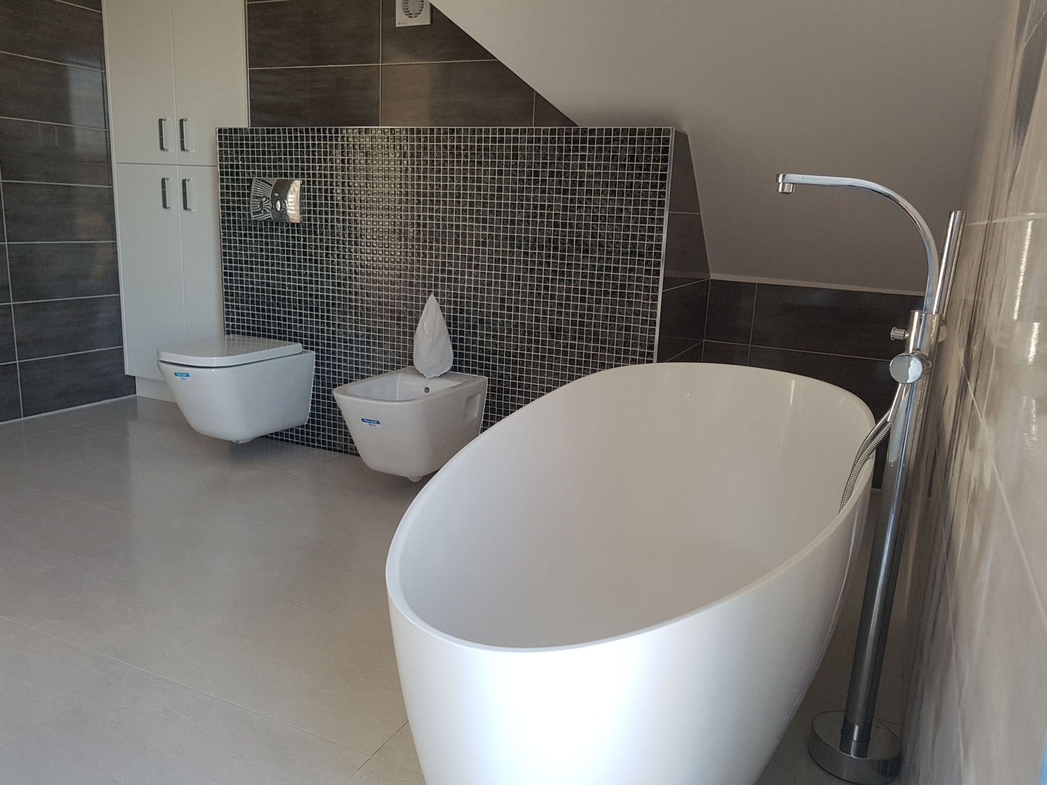 Nowoczesna łazienka na poddaszu z wolnostojącą białą wanną, bidetem i toaletą w stylu minimalistycznym, wykończona szarą mozaiką i płytkami.