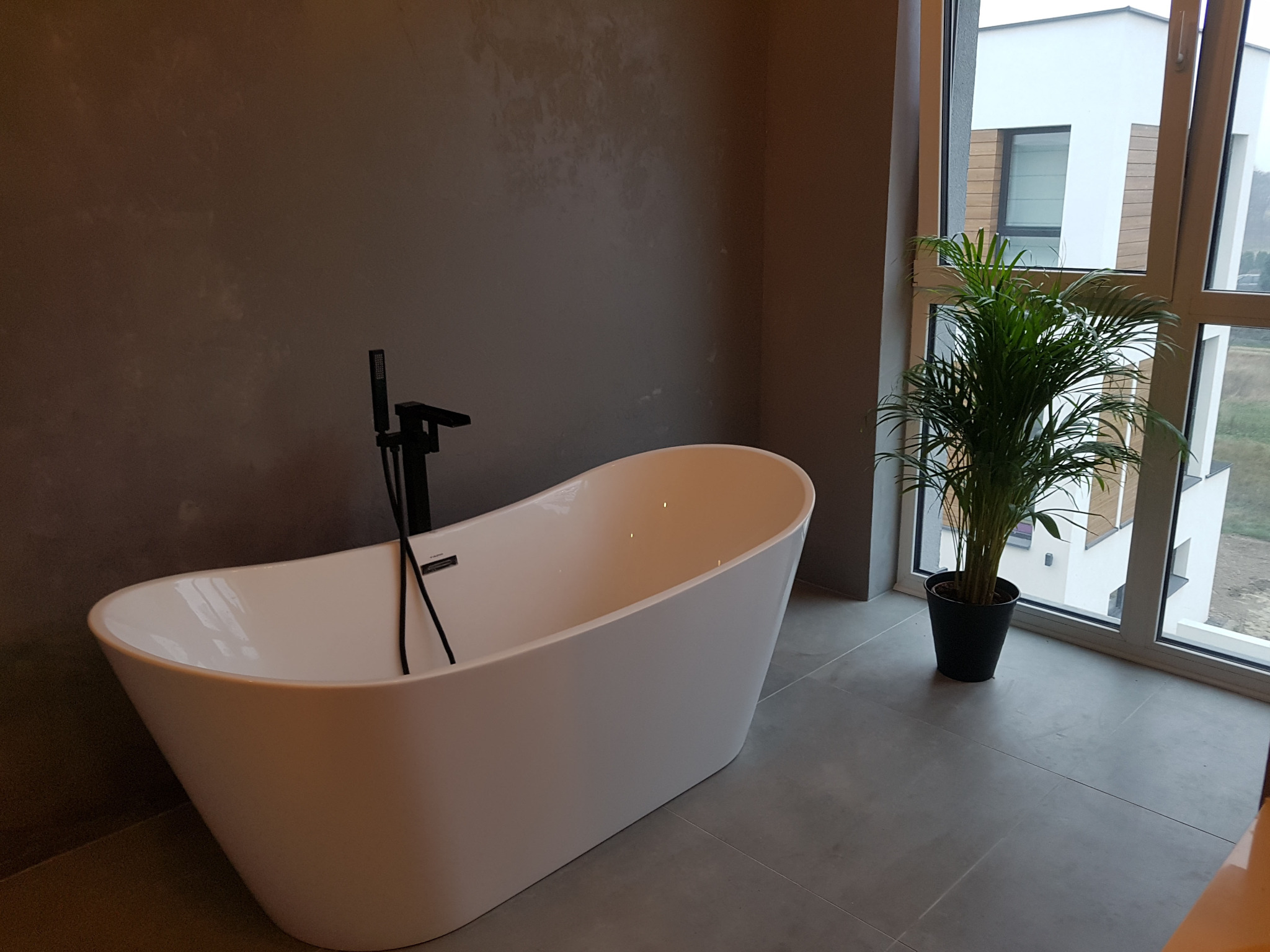 Nowoczesna łazienka z wolnostojącą białą wanną, czarną armaturą i doniczkową palmą przy oknie z widokiem na budynek.