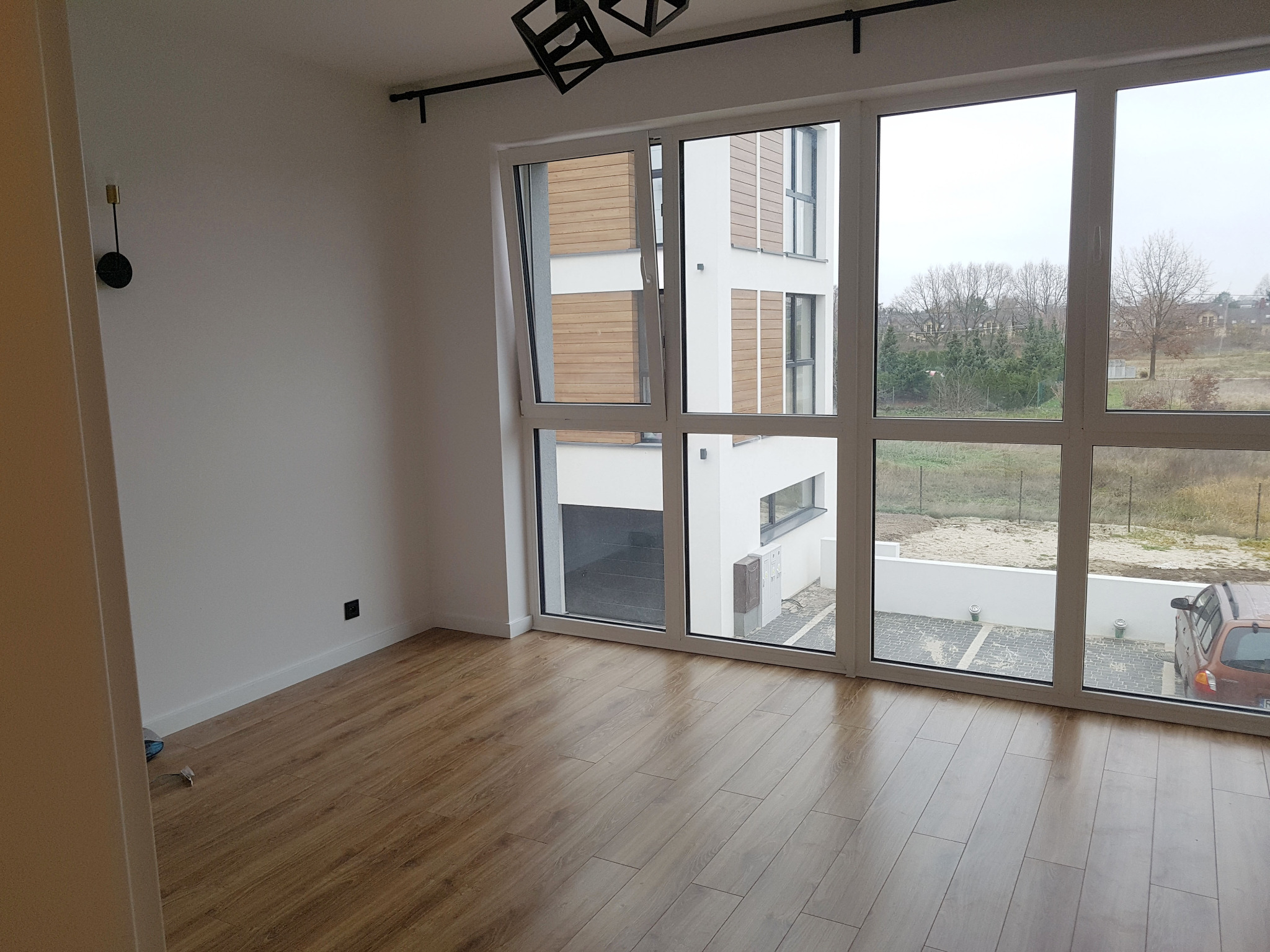 Pusty pokój z panelami podłogowymi w odcieniach brązu, duże okno z widokiem na parking i budynek z drewnianą elewacją, nowoczesna lampa sufitowa z geometrycznym wzorem.