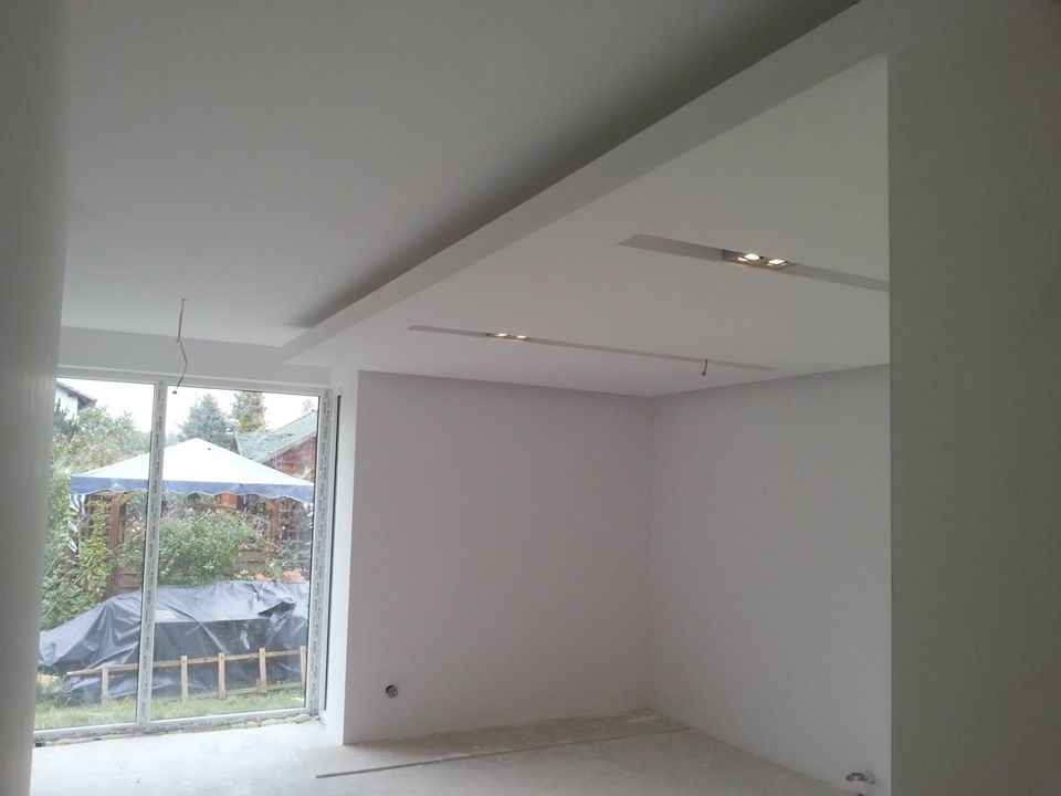 Wnętrze pokoju po malowaniu ścian na biało, widoczne wbudowane oświetlenie sufitowe i duże okno z widokiem na ogród z altaną przykrytą folią.