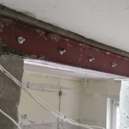 Master-Builders Usługi Wykończeniowe i Budowlane - Wzmocnienie otworu w ścianie za pomocą stalowej belki, widoczne ślady betonu i wystające kable elektryczne, wnętrze w trakcie prac.